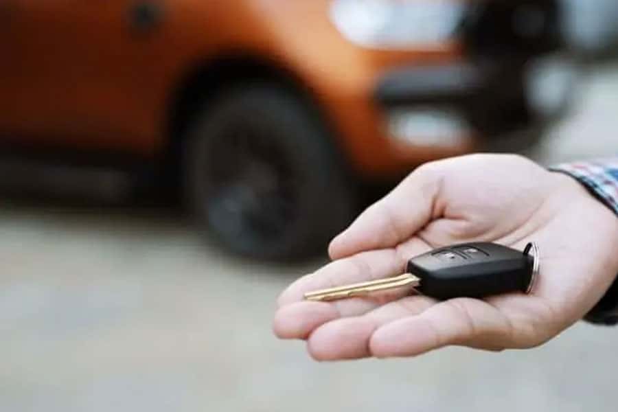 A man extending his hand with a car key in it with the blurred image of an orange car in the background.