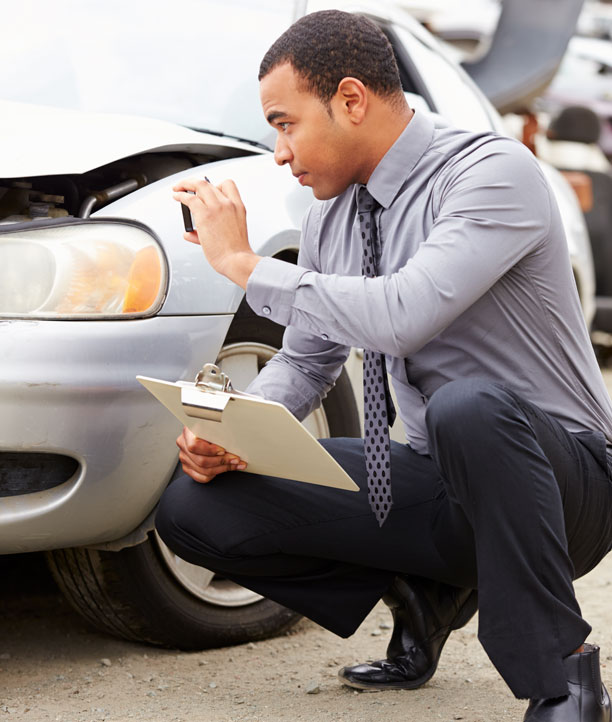 A man wearing a silver dress shirt and black slacks kneels next to a silver car and takes a picture of body damage with his cell phone as he holds a clipboard.